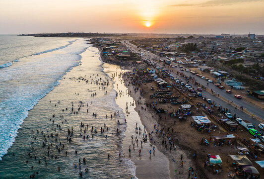 Aerial view of a crowded beach at sunset with people swimming in the ocean, small fishing boats on the sand, and a coastal road with traffic in Ghana.
