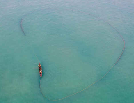 Aerial view of a traditional wooden fishing boat pulling a large circular net through the turquoise waters of the Atlantic Ocean off the coast of Ghana.