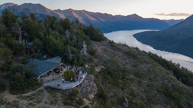 Aerial view of a mountain lodge on a rocky cliff overlooking a lake with people on the terrace at sunset, San Carlos de Bariloche, Rio Negro Province, Argentina.