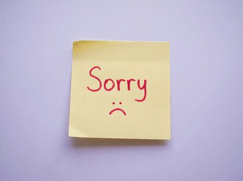 Yellow sticky note with handwritten sorry message and sad face drawing on a light purple background
