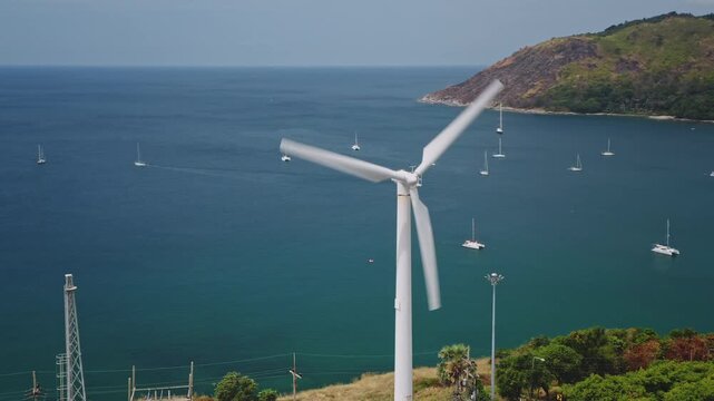 Aerial view panorama showing wind turbine blades spinning, generating renewable energy, overlooking a beautiful bay with numerous sailboats on the turquoise ocean water on Phuket island, Thailand