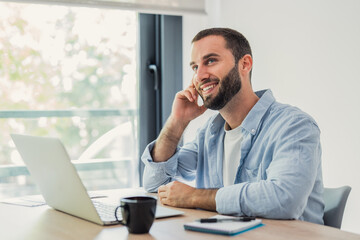 Naklejka premium Smiling bearded man sitting at a desk, looking away and thinking, working on a laptop with a coffee cup and notebook in a bright home office, envisioning future success