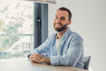 Fototapeta premium Smiling young bearded man sitting at a table near a window. Looking directly at the camera with a happy expression. Portraying positivity. Confidence. And a casual lifestyle in a bright indoor setting