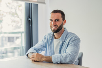 Fototapeta premium A smiling young man in a light blue shirt sits at a wooden desk by a large window, conveying relaxed confidence and approachability in a bright home office or remote work environment.