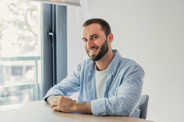 Fototapeta premium Bearded man in a blue shirt and white t-shirt sitting at a wooden table smiling at the camera. Embodying a relaxed. Happy. And confident lifestyle in a bright. Modern indoor setting with natural light