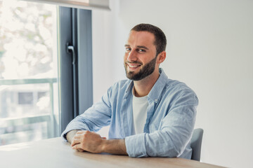 Fototapeta premium Young bearded man sitting at a desk in a modern office, smiling confidently while looking at the camera, portraying a vision of positivity and success in a professional setting