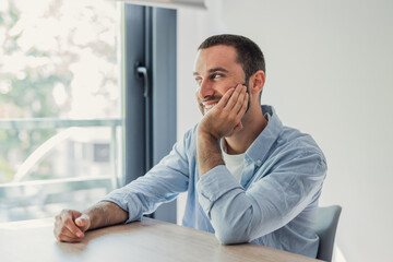 Fototapeta premium Young adult man sitting at a desk, looking out the window with a happy and dreamy expression, resting his chin on his hand