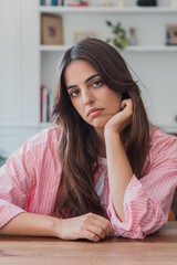Fototapeta premium Young woman sits at a table with her head resting on her hand, appearing pensive, bored and reflective in a casual home interior. Natural, candid portrait conveying mood and contemplation.