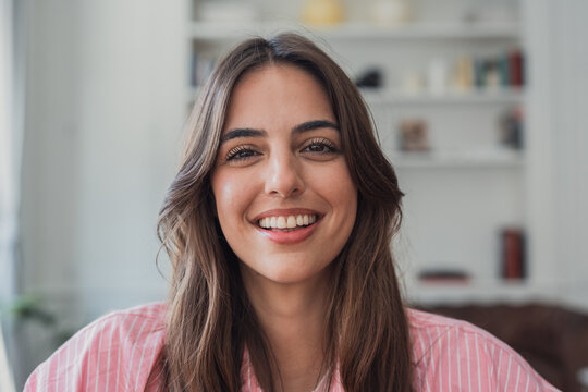 Young brunette woman expresses joy and happiness, smiling broadly at the camera while relaxing at her home office, embodying wellbeing and a positive lifestyle