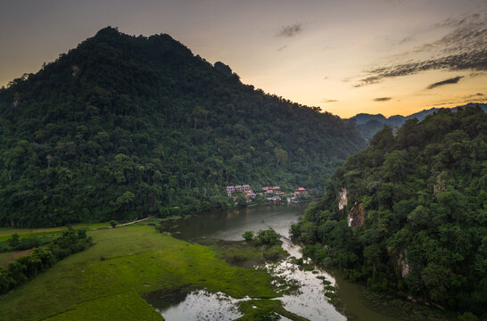 Aerial view of Ba Be Lake surrounded by lush green mountains, calm water, and small houses at sunset in Ba Be, Thai Nguyen, Vietnam.