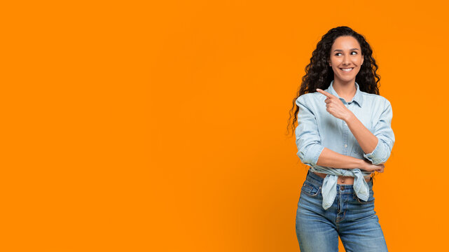 A woman stands with her arms crossed and smiles while pointing to the side against a bright orange backdrop. Her curly hair and casual outfit add a relaxed vibe to the scene
