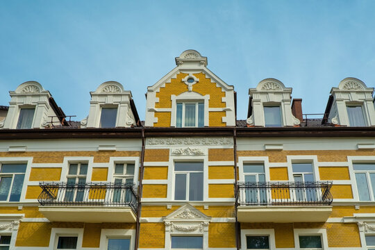 Facade of a historic European building with yellow brick walls and white stucco ornaments. Low angle view of windows, balconies with wrought iron railings, and attic dormers against a clear sky.