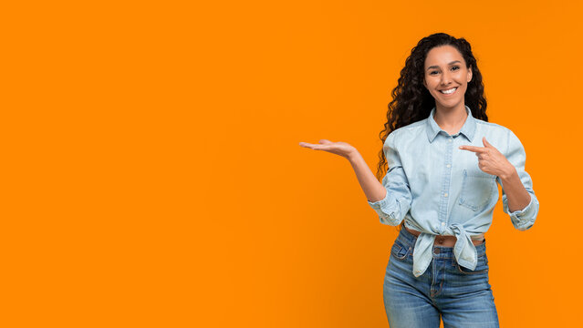 A woman with curly hair smiles while standing in front of an orange wall. She points with one hand and holds the other hand open. The setting is bright and simple, focusing on her playful pose