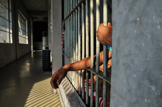 A man in prison holds the steel bars of the cell with his hands behind his back; a criminal locked up in jail.
