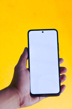 Vertical photo of a person's hand holding a smartphone with a glowing blank white screen for app mockup against a bright yellow background.