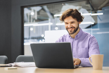 Fototapeta premium A man sits at a table in a modern office, using a laptop. He appears focused and is smiling, enjoying his work. A coffee cup is nearby on the table