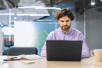 A man sits at a desk in an office, focused on his laptop. He has a serious expression as he types....