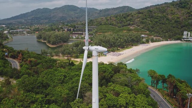 Wind turbine blades spinning, creating renewable energy on a lush green hillside overlooking turquoise bay with yachts anchored, pristine sandy beach, luxury resorts in Phuket, Thailand. Aerial shot