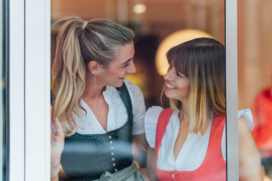 Portrait of two women dressed in traditional Bavarian dirndl costumes looking into a Tracht shop window in the heart of Bavaria.