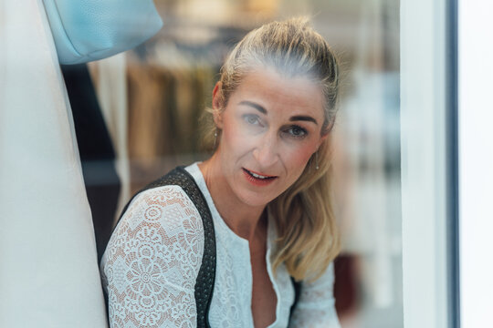 Portrait of a smiling blonde woman in a traditional Bavarian dirndl looking through the window of a specialty Tracht clothing store.
