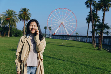 Obraz premium Smiling woman talking on phone in a sunny park, ferris wheel in the distance, green grass, palm trees, casual coat, daytime outdoor scene for communication, travel planning and leisure
