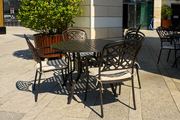 Fototapeta premium Empty patio furniture set featuring black forged iron openwork table and chairs with cushions standing on sunny city terrace.