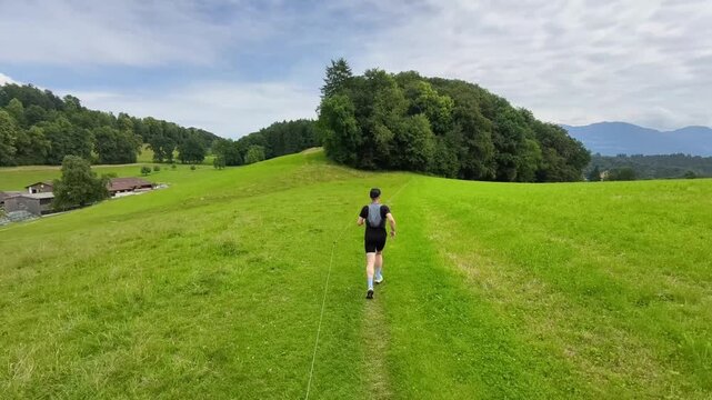 A trail runner follows a narrow path across rolling green meadows surrounded by forests and rural farmland in Switzerland. Ideal for concepts of fitness, adventure, and outdoor recreation.