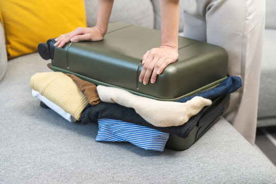 Full suitcase concept, hand of asian young woman, girl packing, preparing luggage case, luggage of clothes, female unable to close bag while sitting on sofa at home, travel trip or journey voyage.
