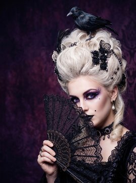 Woman in dark gothic baroque costume with blonde wig decorated with black roses and skulls, heavy purple smoky makeup, holding black lace spiderweb fan against purple background.