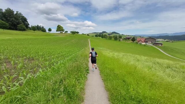 A trail runner follows a narrow path across rolling green meadows surrounded by forests and rural farmland in Switzerland. Ideal for concepts of fitness, adventure, and outdoor recreation.