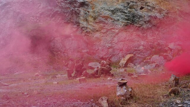 A multinational group of male soldiers in infantry uniforms, carrying paintball guns and ready for combat, were seen running and practicing military drills, with pink smoke signals being sent 