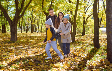 Obraz premium Family with children at play in autumn park. Parents lead kids in a playful line through leaves, laughing together amid sunny nature on a carefree weekend. Happy, warm family bonding in fall.