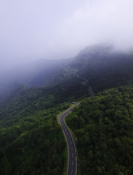 Aerial view of a winding mountain road cutting through lush green forests and misty hills in the Western Ghats near Karad, Maharashtra, India.