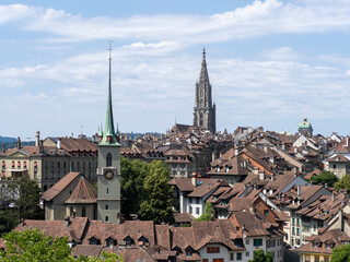 Fototapeta premium Panoramic view over the red-tiled rooftops of Bern's UNESCO World Heritage old town with the green spire of the Nydegg church and the Gothic tower of Bern Minster under a blue summer sky, Switzerland