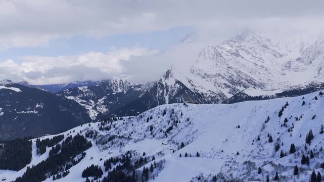 Wide aerial panoramic footage of the French Alps in winter, showing snow-covered mountains, forested slopes, and open alpine terrain. The scene includes rolling snowy hills, scattered evergreen
