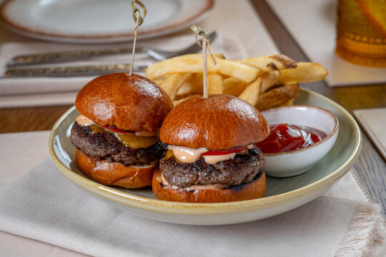 Delicious mini burgers with fries served in a casual restaurant setting. Two mini burgers with toppings and fries are on a plate in a restaurant.
