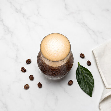 Overhead Flat Lay of Nitro Cold Brew Coffee in Glass with Creamy Foam on White Marble, Coffee Beans and Fresh Leaf Scattered Around, Minimal Aerial View Food Photography