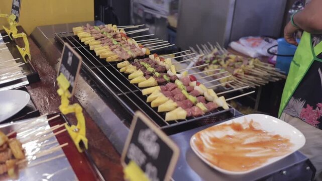 Thai BBQ Street Food Vendor Grilling Cooks Pork Meat Skewer Kebabs, with Pineapples Vegetables, Brushes Sauce. Asian Bangkok Thailand Open Night Market