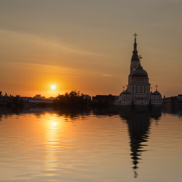 The sun sets behind a tall church with domes and crosses. The sky shows warm colors and the water reflects the building. It is evening near a city with a calm atmosphere.