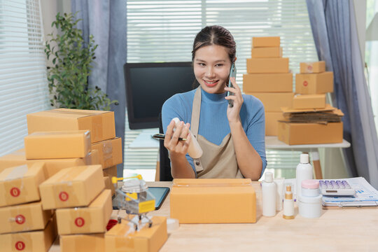 Smiling young Asian woman online seller packaging cosmetics skincare products into cardboard parcel boxes at home office, E-commerce small business owner concept.