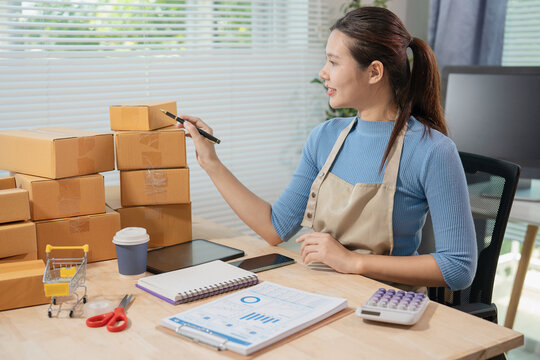 Smiling young Asian woman online seller packaging cosmetics skincare products into cardboard parcel boxes at home office, E-commerce small business owner concept.
