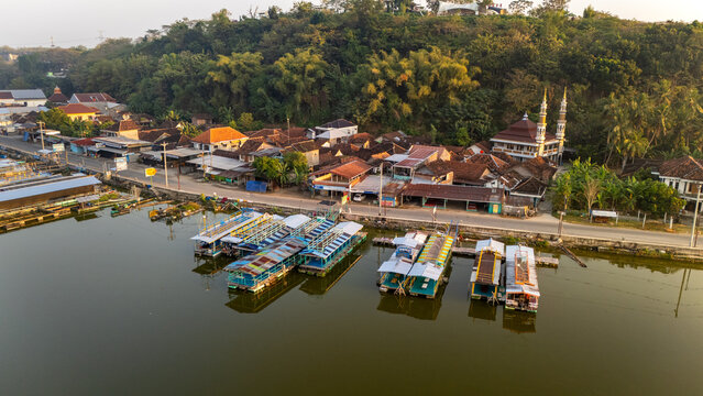 Aerial view of a lakeside village in Indonesia featuring floating boats moored along the shore, a mosque with twin minarets, and lush green hills in the background, Indonesia.