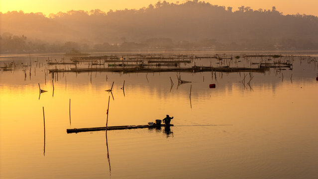 Aerial view of a lone fisherman on a bamboo raft surrounded by traditional fish traps in a calm lake during golden hour in Indonesia.
