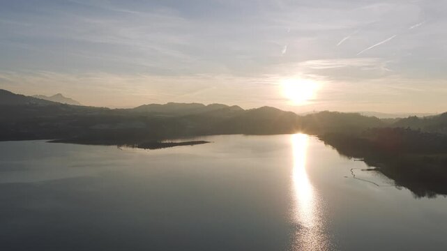 Aerial footage captures the tranquil beauty of Lake Aiguebelette at sunrise in Savoie, France. The calm water reflects the soft morning light, with distant hills and a small island visible. The sky