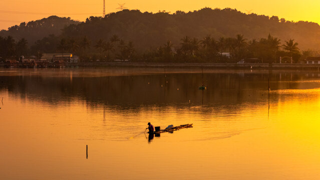 Aerial view of a lone fisherman on a small bamboo raft in a calm lake during golden hour with lush hills in the background in Indonesia.