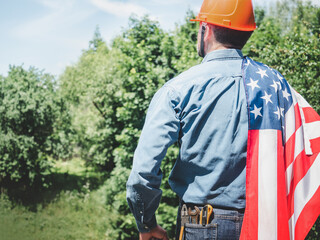 Obraz premium Construction worker in an orange hard hat with an American flag draped over his shoulder. Symbolizes American workers or Labor Day. Concept of pride in construction profession and American dream