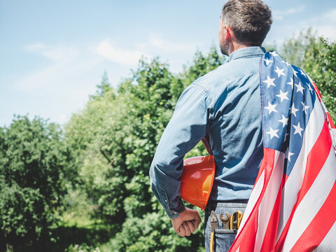 Construction worker in an orange hard hat with an American flag draped over his shoulder. Symbolizes American workers or Labor Day. Concept of pride in construction profession and American dream