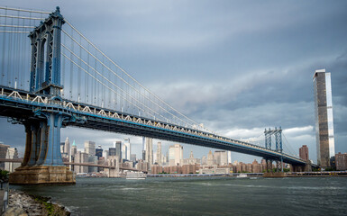 Obraz premium New York, USA, 15 June 2026: View beneath Manhattan Bridge, New York City. Angled river perspective with suspension cables, skyline backdrop and soft daylight.