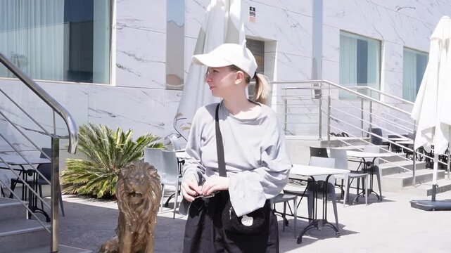 A young woman wearing a youth cap with earflaps smiles, poses, and presents sunglasses near a cafe terrace on a hot day. Leisure accessories. Youthful clothing style.