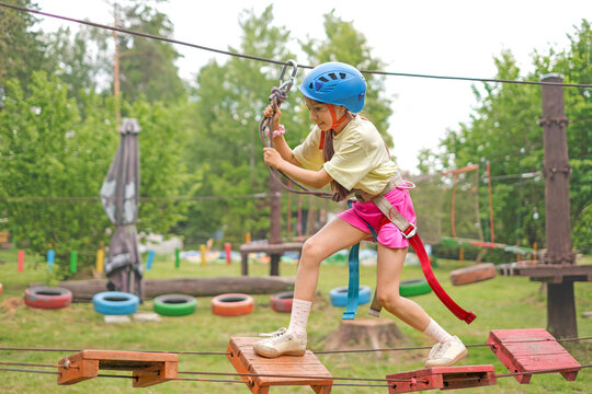 A girl wearing protective sports gear and a helmet is walking along a suspended bridge. Outdoor recreation and entertainment park
